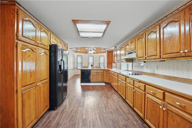 kitchen with brown cabinets, a peninsula, dark wood-type flooring, under cabinet range hood, and black appliances