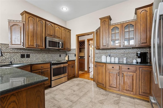 kitchen with backsplash, appliances with stainless steel finishes, a sink, dark stone countertops, and glass insert cabinets