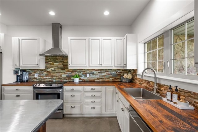 kitchen featuring butcher block countertops, wall chimney range hood, stainless steel appliances, a sink, and white cabinetry