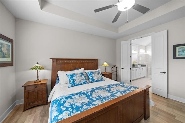 bedroom featuring light wood-style floors, baseboards, a tray ceiling, ensuite bathroom, and ceiling fan