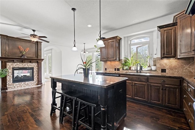 kitchen with a fireplace, dark stone countertops, dark wood-style floors, a sink, and decorative backsplash