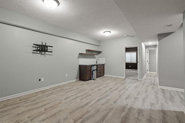basement featuring a textured ceiling, light wood-type flooring, baseboards, and bar with sink