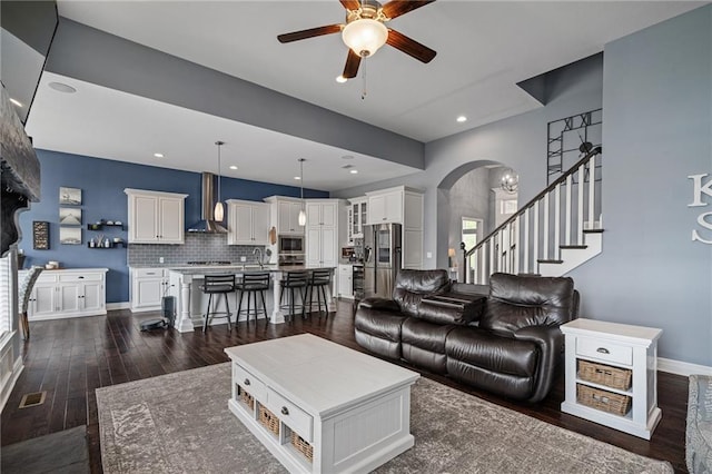 living room with a ceiling fan, arched walkways, stairway, baseboards, and dark wood-style floors