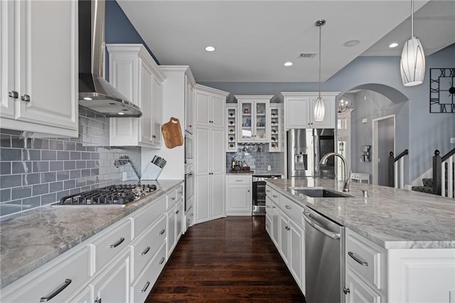 kitchen with appliances with stainless steel finishes, a sink, arched walkways, wall chimney exhaust hood, and tasteful backsplash