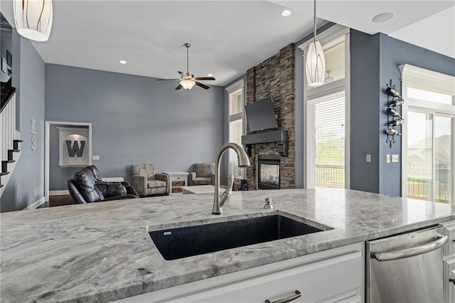 kitchen featuring ceiling fan, light stone countertops, a sink, dishwasher, and a stone fireplace