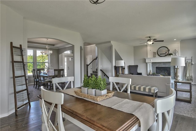 dining room featuring stairway, a ceiling fan, arched walkways, a glass covered fireplace, and dark wood-style flooring