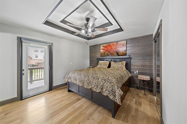 bedroom featuring access to outside, ceiling fan, light wood-type flooring, wooden walls, and a tray ceiling