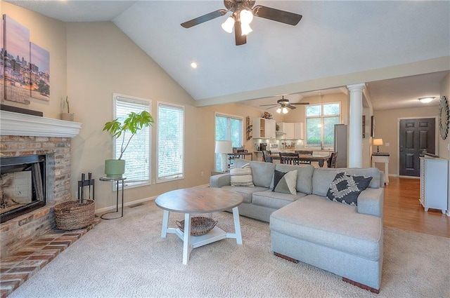 carpeted living room with ceiling fan, baseboards, a brick fireplace, high vaulted ceiling, and decorative columns