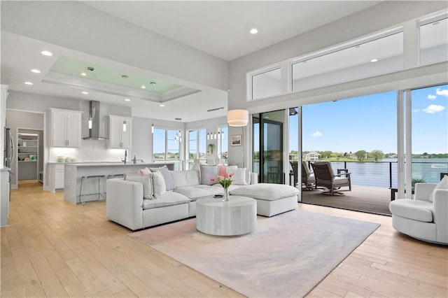 living room with light wood-type flooring, a raised ceiling, a water view, and recessed lighting