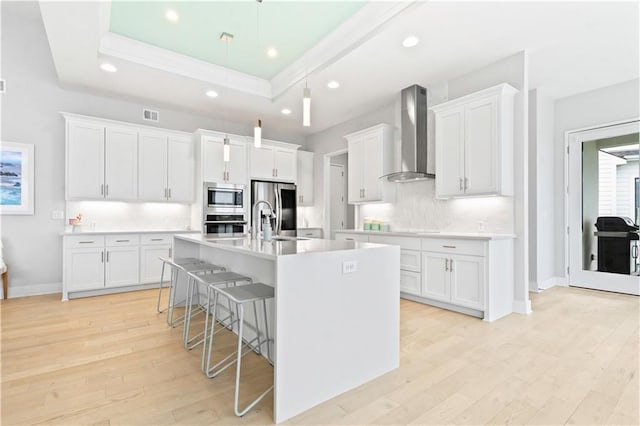kitchen with wall chimney range hood, appliances with stainless steel finishes, a tray ceiling, a kitchen bar, and light wood-type flooring