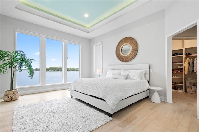 bedroom with light wood-style flooring, a spacious closet, a tray ceiling, a water view, and recessed lighting