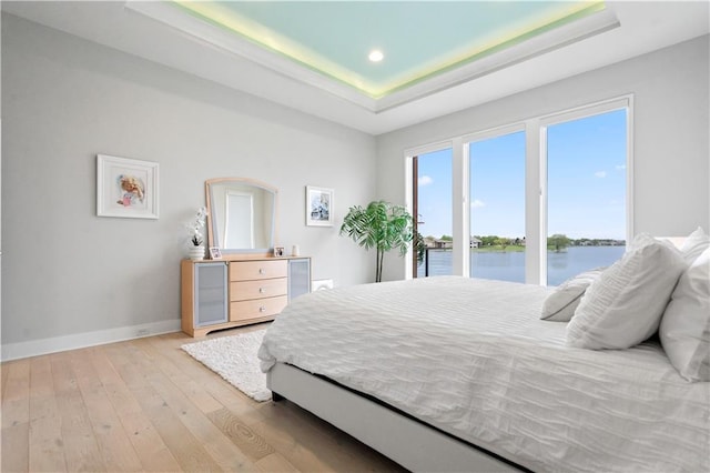 bedroom with a tray ceiling, baseboards, a water view, wood-type flooring, and access to outside