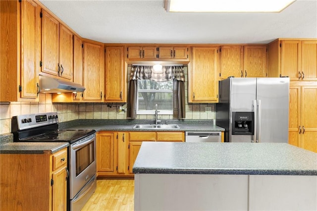 kitchen with light wood finished floors, a sink, under cabinet range hood, dark countertops, and appliances with stainless steel finishes