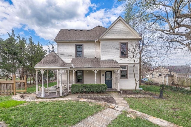 victorian-style house featuring a front yard, roof with shingles, covered porch, and fence
