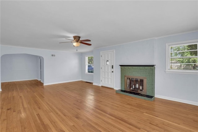 unfurnished living room with ceiling fan, a brick fireplace, arched walkways, baseboards, and light wood-type flooring