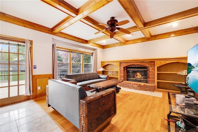 living room featuring ceiling fan, a fireplace, a wainscoted wall, beamed ceiling, and light wood-style floors