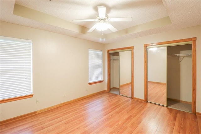 unfurnished bedroom featuring two closets, a raised ceiling, light wood-style flooring, baseboards, and a textured ceiling
