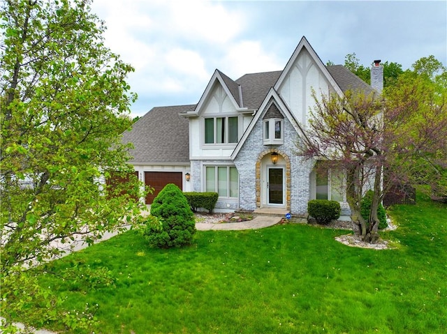 tudor house with a chimney, stucco siding, roof with shingles, a front yard, and an attached garage