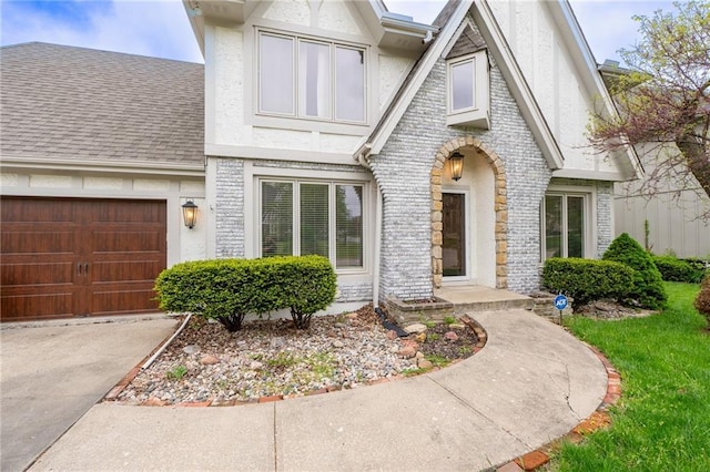 tudor-style house with concrete driveway, a shingled roof, stucco siding, brick siding, and a garage