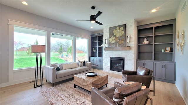 living room with light wood-style floors, baseboards, a ceiling fan, built in features, and a stone fireplace