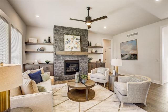 living area featuring a stone fireplace, light wood-style flooring, a ceiling fan, baseboards, and visible vents
