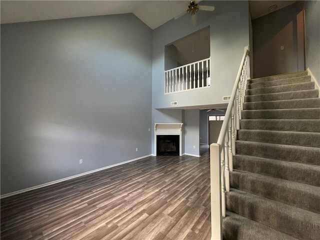 staircase featuring a ceiling fan, a fireplace, wood finished floors, high vaulted ceiling, and baseboards