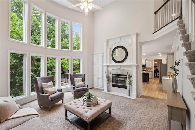 living room featuring a towering ceiling, healthy amount of natural light, a ceiling fan, a tiled fireplace, and light carpet