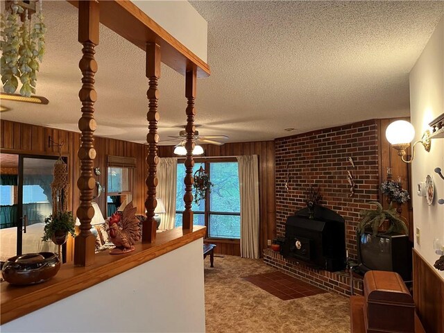 carpeted living area with wooden walls, ceiling fan, a wood stove, and a textured ceiling