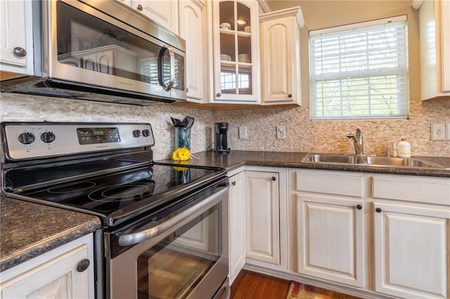 kitchen with dark countertops, a sink, stainless steel appliances, backsplash, and glass insert cabinets