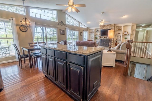 kitchen with dark wood-style flooring, light stone countertops, ceiling fan with notable chandelier, a center island, and lofted ceiling