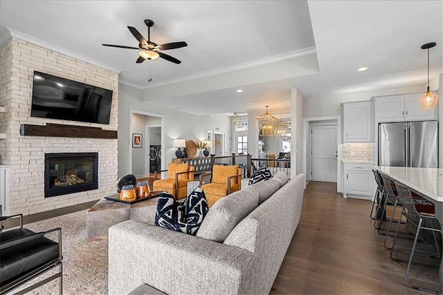 living area featuring ornamental molding, dark wood-type flooring, recessed lighting, a large fireplace, and ceiling fan with notable chandelier