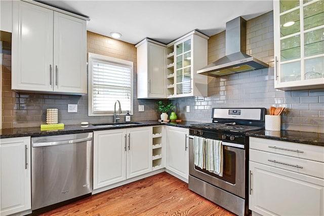 kitchen featuring open shelves, wall chimney range hood, light wood-type flooring, a sink, and stainless steel appliances