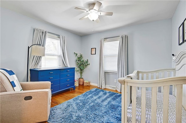 bedroom featuring a nursery area, ceiling fan, baseboards, and wood finished floors