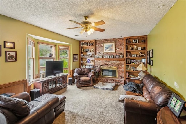 carpeted living room featuring ceiling fan, a textured ceiling, built in features, a brick fireplace, and a wainscoted wall