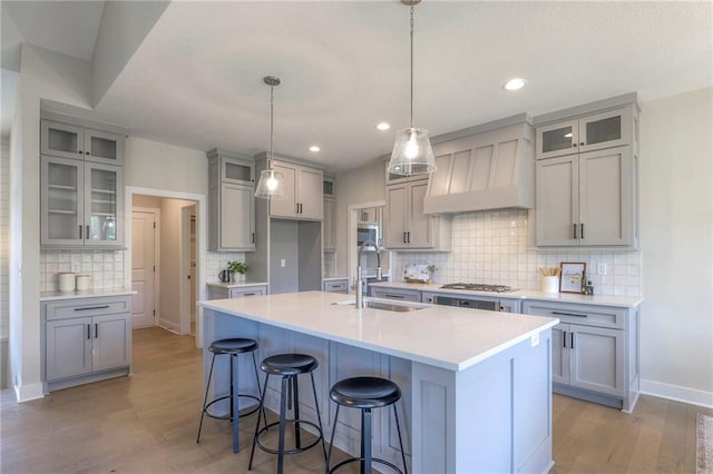 kitchen featuring backsplash, a center island with sink, a sink, premium range hood, and light wood finished floors
