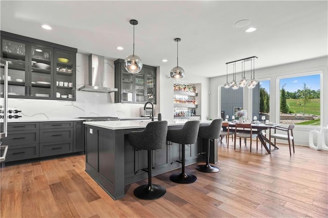 kitchen with wall chimney range hood, a breakfast bar, light countertops, light wood-type flooring, and glass insert cabinets