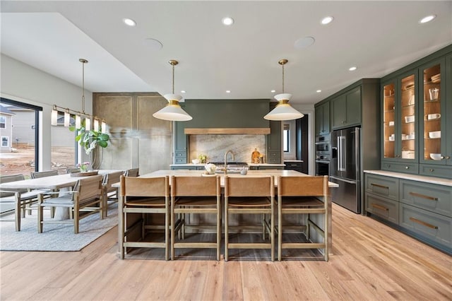 kitchen featuring high end fridge, a center island with sink, light wood-type flooring, light countertops, and hanging light fixtures