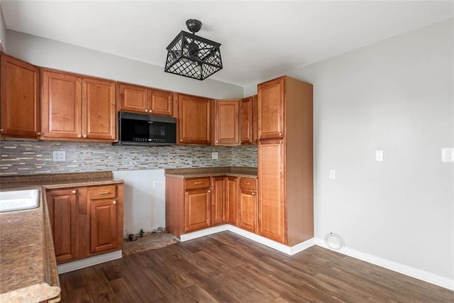 kitchen featuring black microwave, dark wood-style flooring, backsplash, brown cabinetry, and baseboards