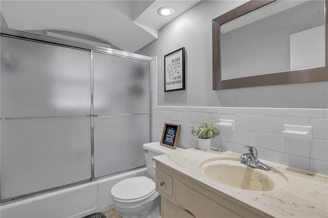 bathroom with toilet, bath / shower combo with glass door, vanity, tile walls, and wainscoting
