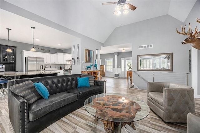 living room with ceiling fan, high vaulted ceiling, light wood-style flooring, decorative columns, and visible vents