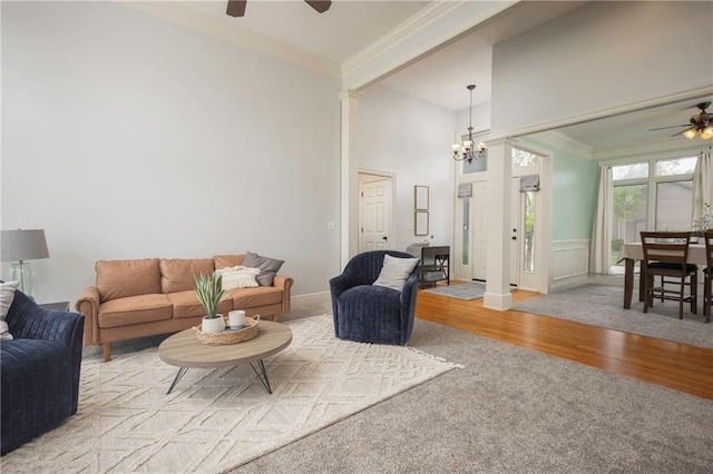 carpeted living room featuring crown molding, a high ceiling, wainscoting, ceiling fan with notable chandelier, and wood finished floors