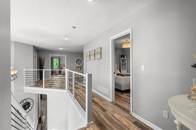 corridor with a textured ceiling, visible vents, wood finished floors, baseboards, and an upstairs landing