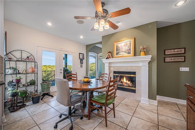 dining space featuring arched walkways, a tiled fireplace, french doors, a ceiling fan, and light tile patterned floors