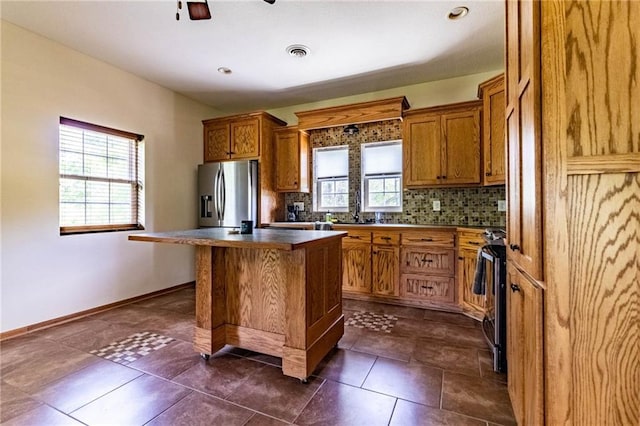 kitchen with appliances with stainless steel finishes, brown cabinetry, backsplash, a center island, and ceiling fan