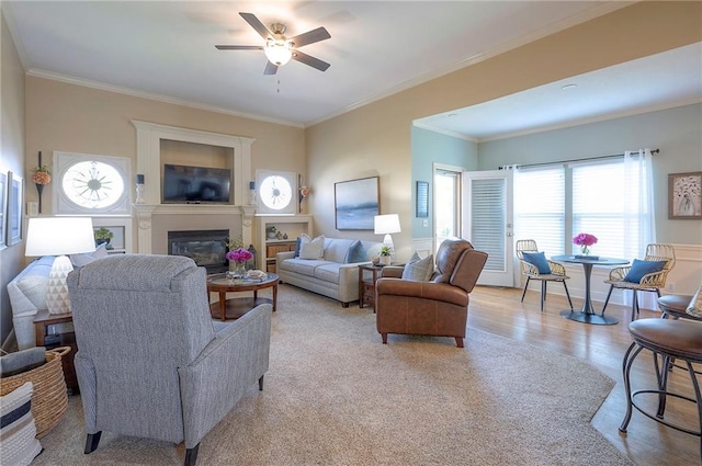 living room with crown molding, light wood-style flooring, a ceiling fan, and a glass covered fireplace