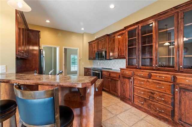 kitchen featuring a kitchen breakfast bar, backsplash, appliances with stainless steel finishes, glass insert cabinets, and light tile patterned flooring