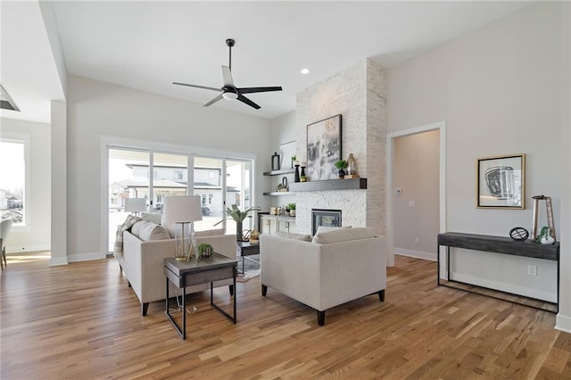 living room featuring a stone fireplace, light wood-type flooring, baseboards, and ceiling fan