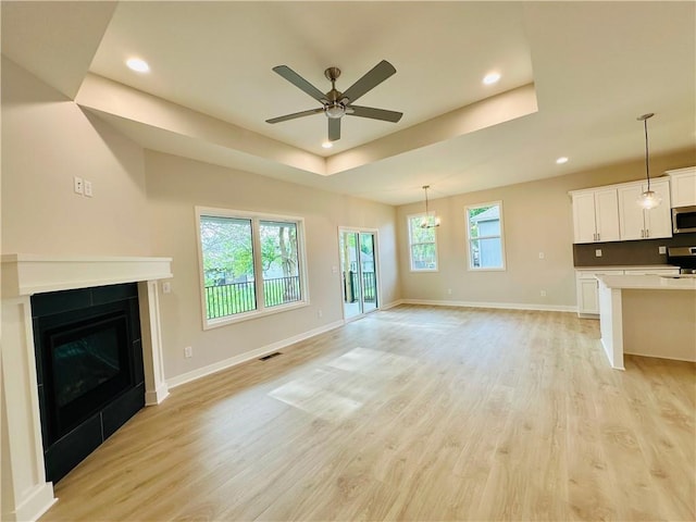 unfurnished living room featuring ceiling fan with notable chandelier, a tray ceiling, recessed lighting, and light wood-style flooring