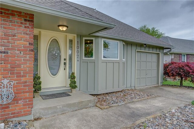 property entrance with roof with shingles, an attached garage, brick siding, board and batten siding, and driveway