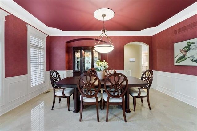 dining room featuring arched walkways, light tile patterned flooring, a decorative wall, crown molding, and wainscoting